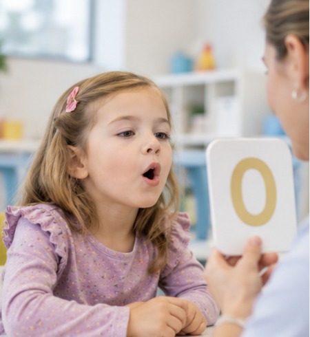 Preschool girl practicing an O sound as a speech therapist holds up a letter card